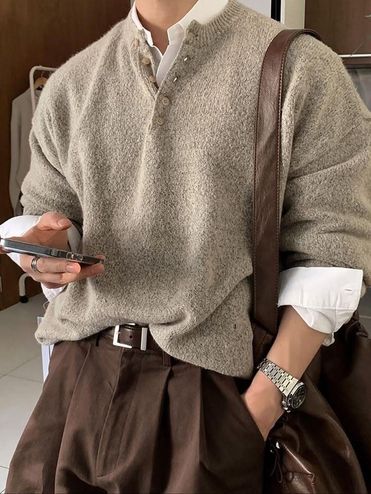 Man wearing a cream knit henley sweater with dark brown pleated trousers and a leather bag.