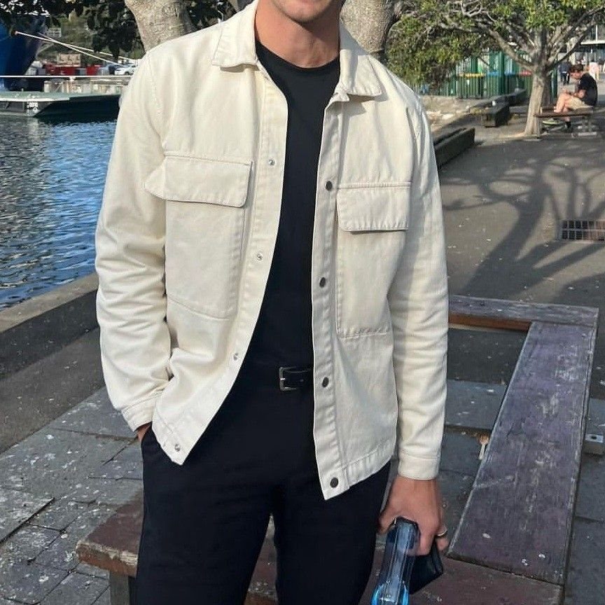 Man wearing a cream button-up jacket over a black t-shirt and black pants outdoors.
