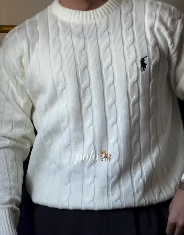 A man wearing a cream cable-knit sweater with a vertical braided texture and small logo.
