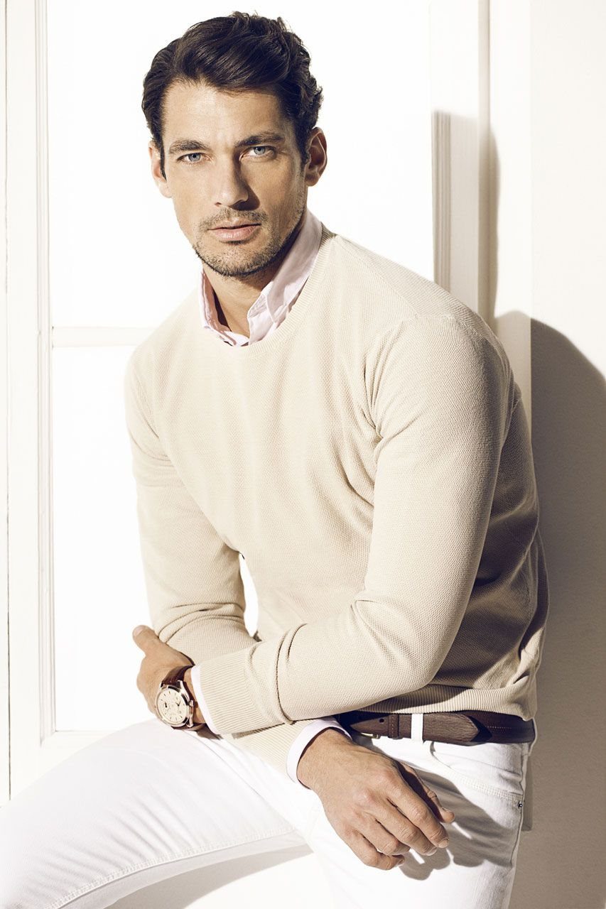 A man posing in a cream knit sweater, pink collared shirt, and white trousers near a window.