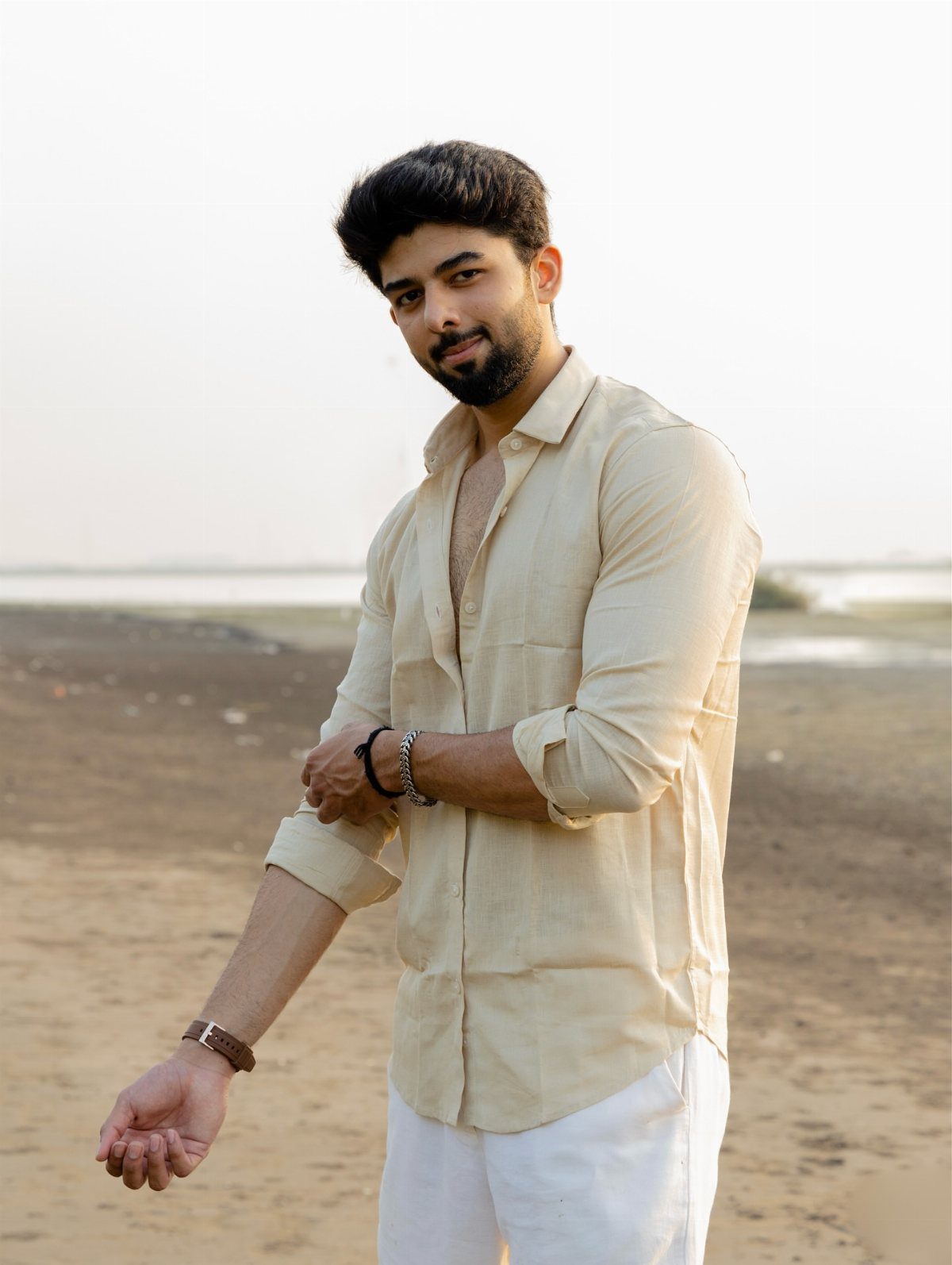 A man wearing a cream linen shirt and white trousers standing on a beach.