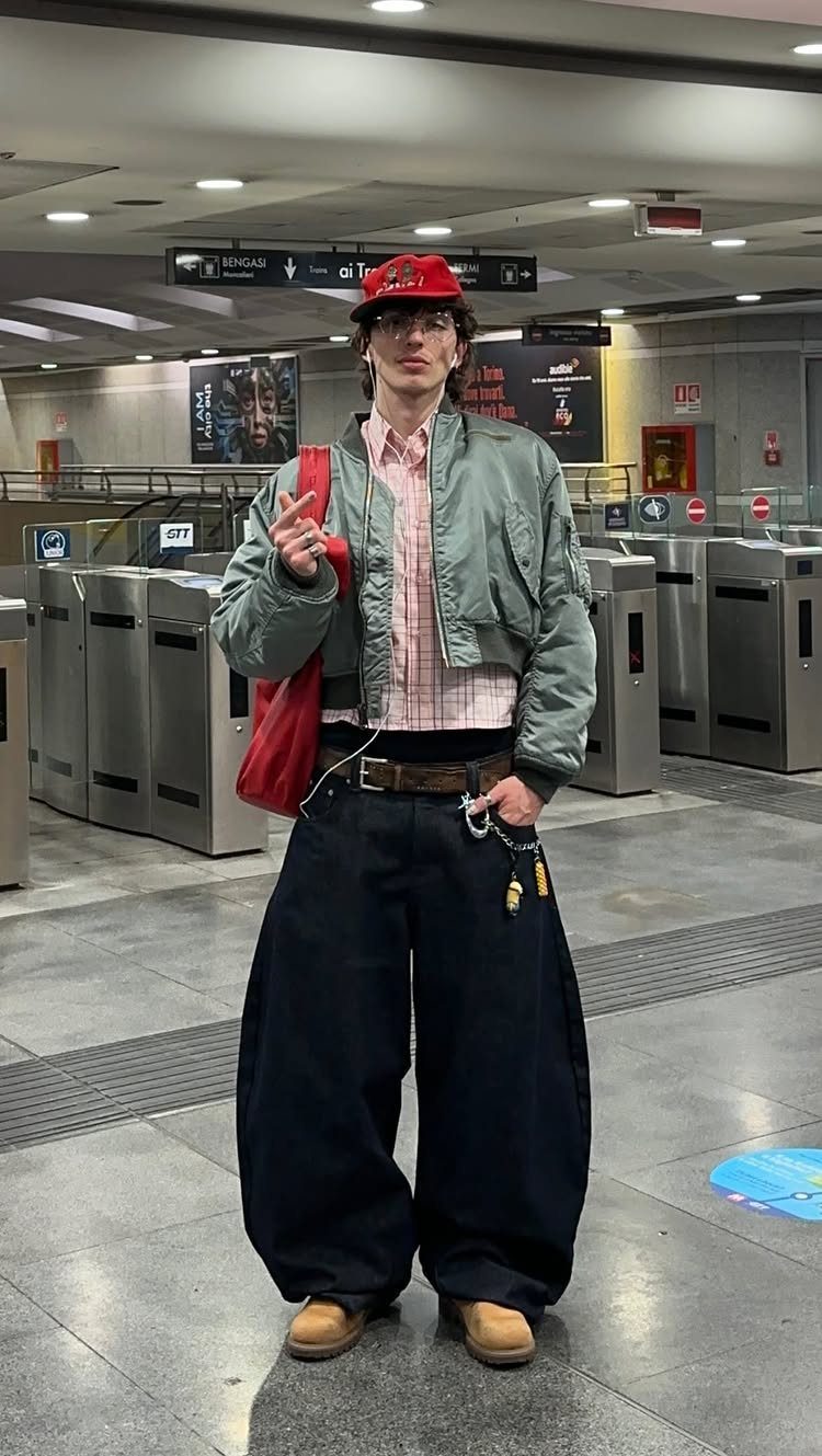 Person wearing a light green cropped bomber jacket, pink checkered shirt, and wide-leg dark pants in a subway station.