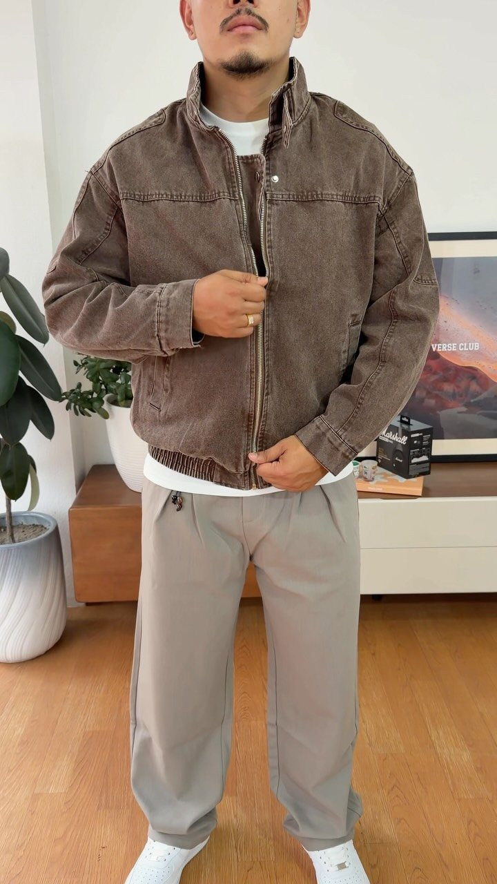 Man wearing a brown denim jacket, white t-shirt, and grey trousers standing indoors.