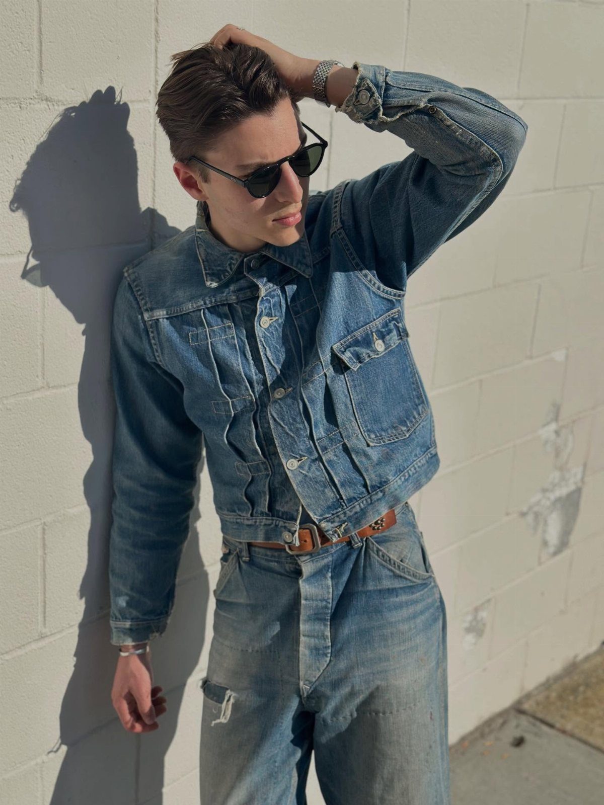 Man in a denim jacket and matching jeans leaning against a white brick wall.