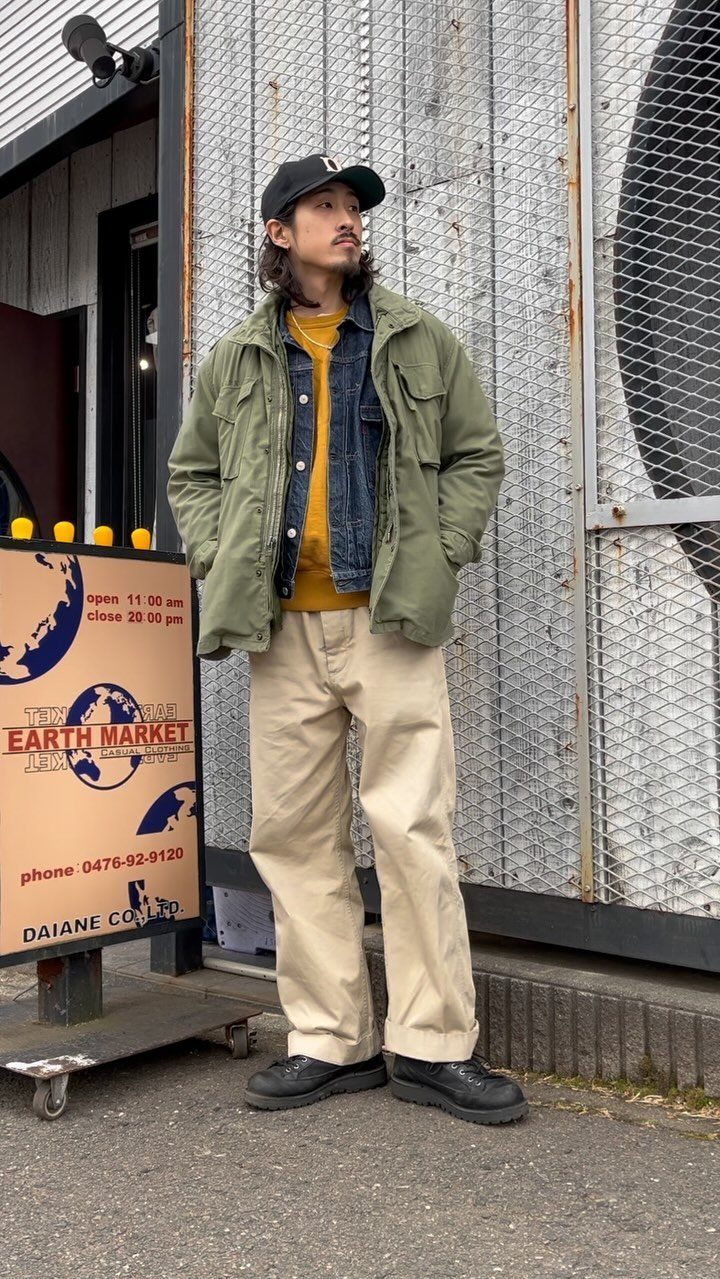 Man wearing a denim jacket, olive utility jacket, yellow shirt, and beige trousers standing outdoors.