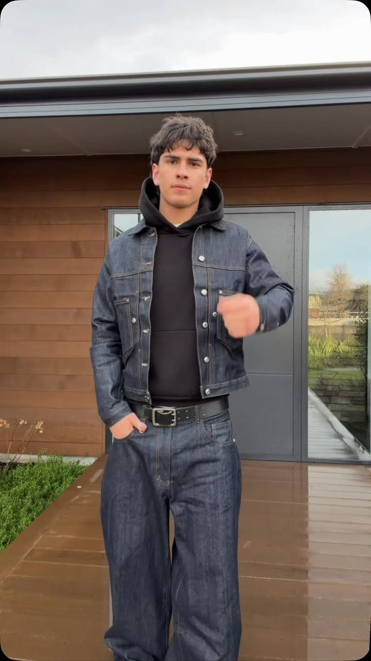 Man wearing a dark denim jacket, black hoodie, and matching wide-leg denim jeans standing outdoors.
