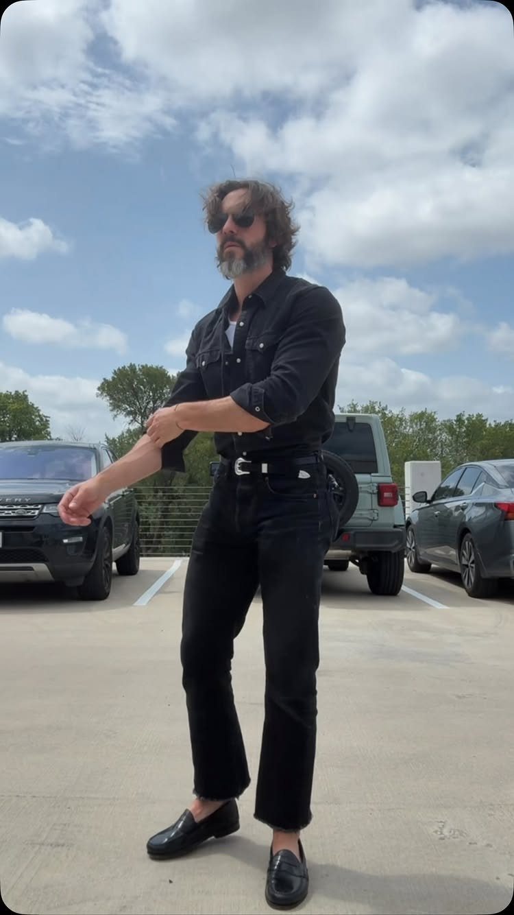 A bearded man in a black denim outfit posing in a parking lot under a cloudy sky.