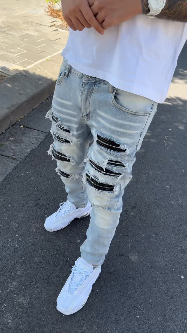 Man wearing light blue distressed denim jeans with black patches and white sneakers on a sidewalk.