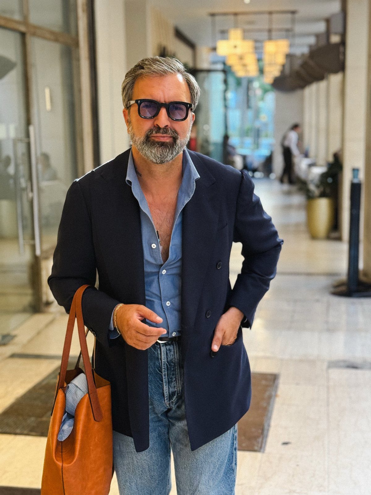 Man wearing a navy blazer, light blue shirt, and denim jeans while carrying a leather bag.
