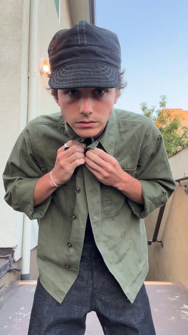 Man in olive green shirt and dark denim jeans wearing a black cadet cap.