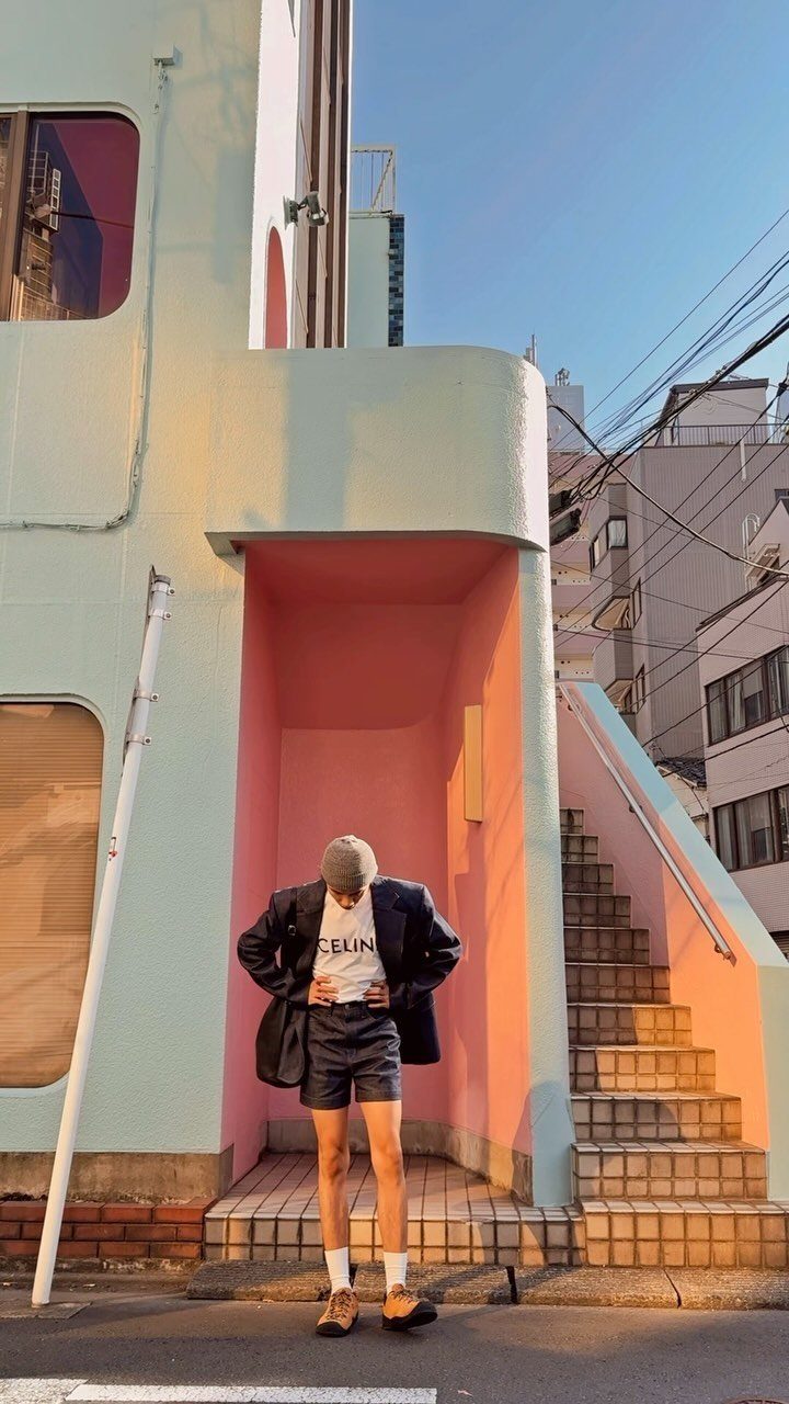Person wearing a black blazer, white graphic tee, and denim shorts standing in a pink alcove.
