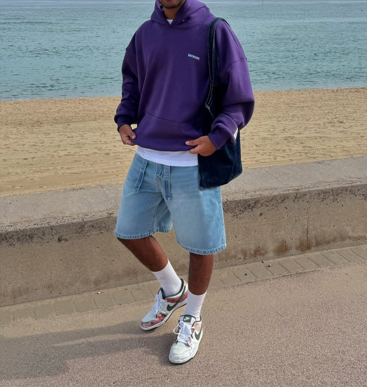 Person wearing a purple hoodie and light blue denim shorts standing near a beach.