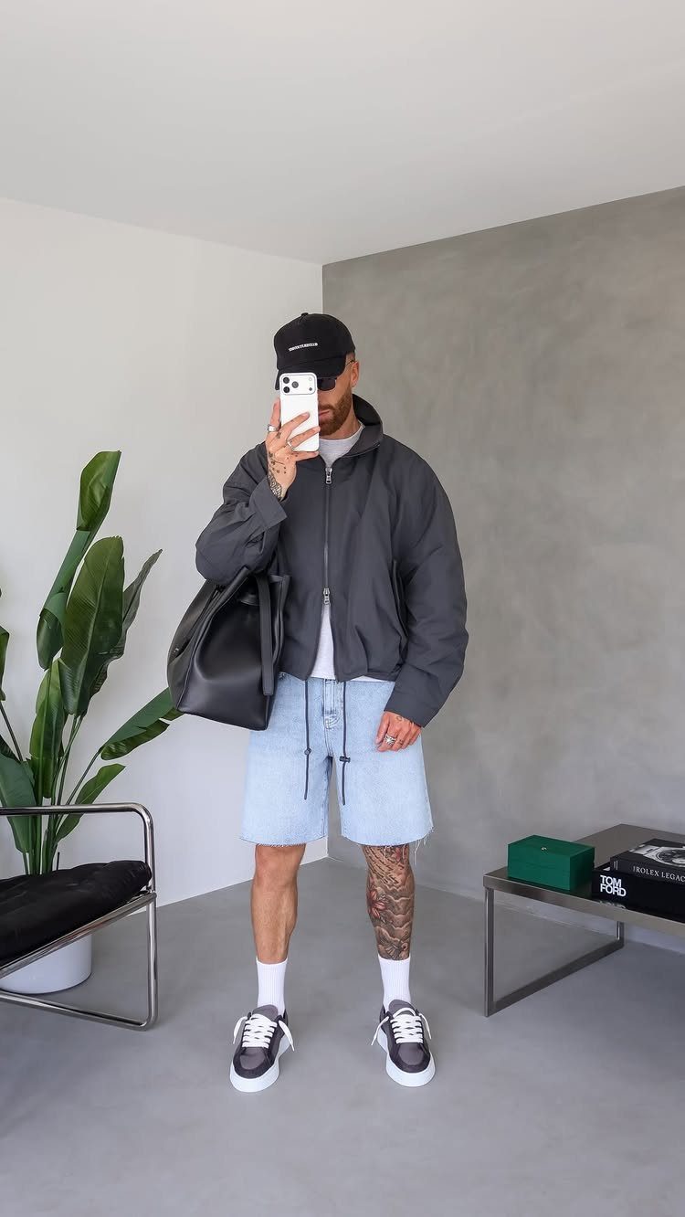 Man in light blue denim shorts, grey jacket, and black cap taking a mirror selfie.