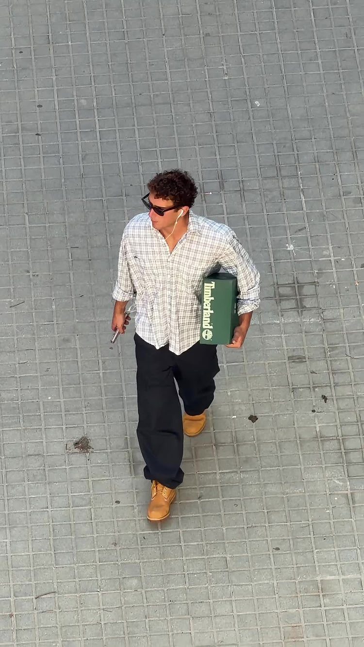 A man in a checkered shirt and dark trousers walking while wearing tan boots.