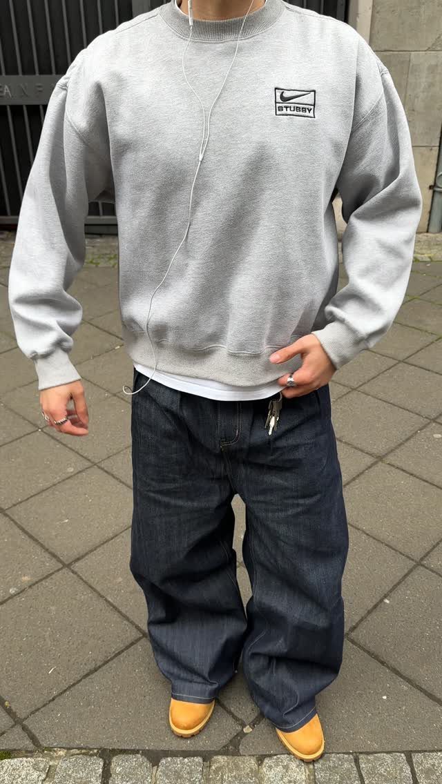 Person wearing a grey sweatshirt, wide-leg dark denim jeans, and tan shoes standing on a sidewalk.