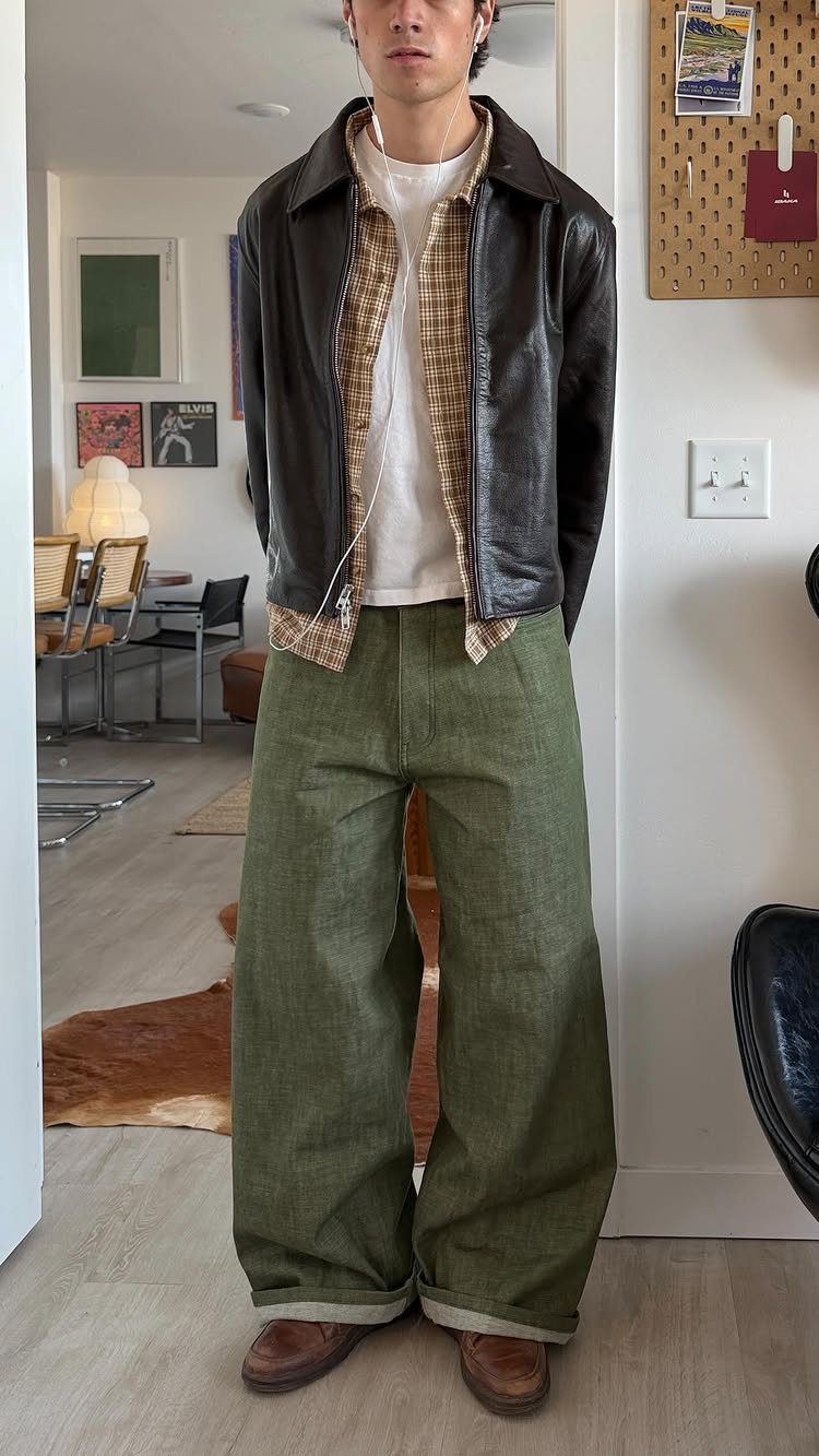 A man wearing a leather jacket, patterned shirt, white t-shirt, and wide olive green trousers.