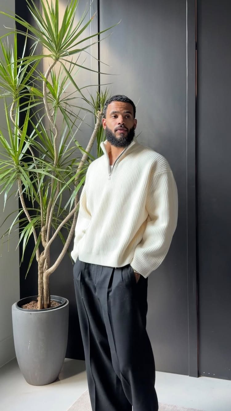 Man in a cream textured sweater and black trousers standing by a large potted plant.