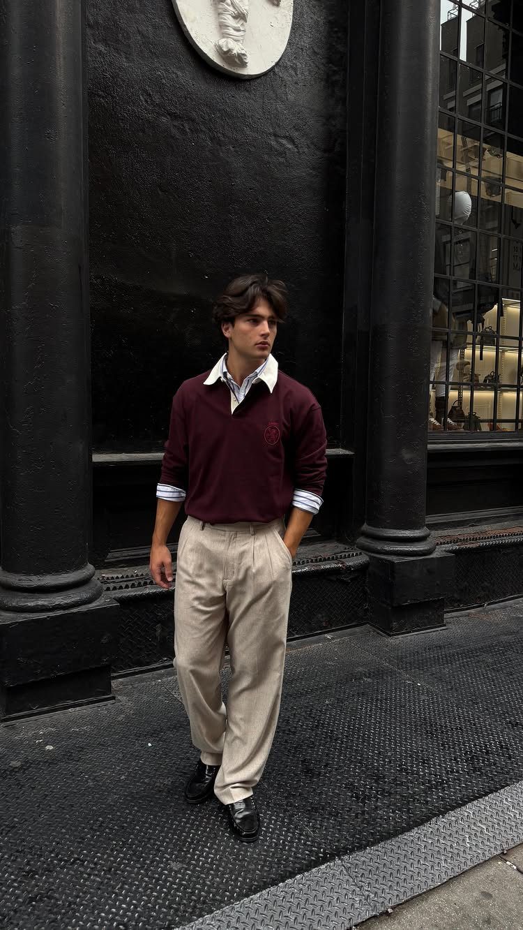 A man wearing a maroon sweater, white collared shirt, and beige trousers standing outdoors.