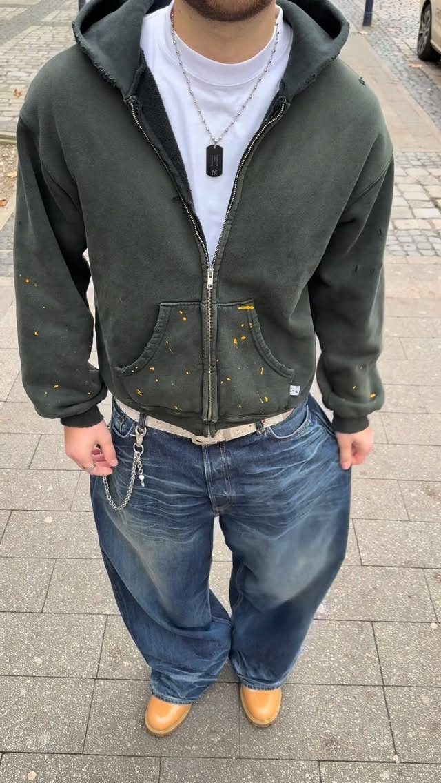 A man wearing a dark green hoodie, wide blue jeans, and tan shoes standing on a sidewalk.