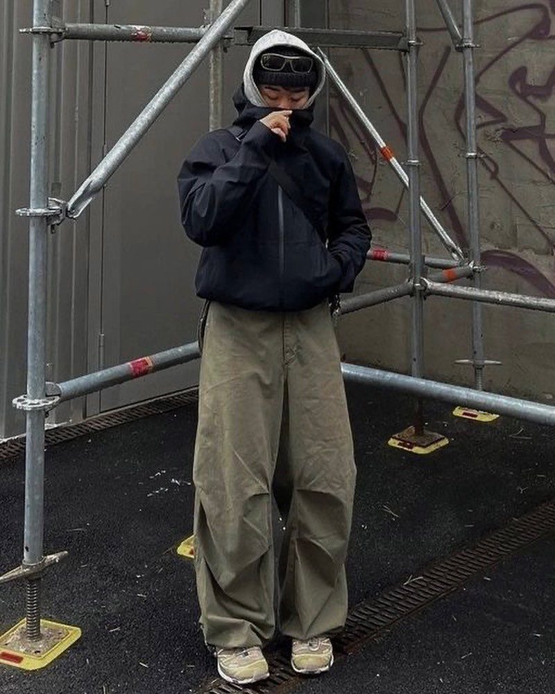 Man in black technical jacket and olive cargo pants standing by metal scaffolding.