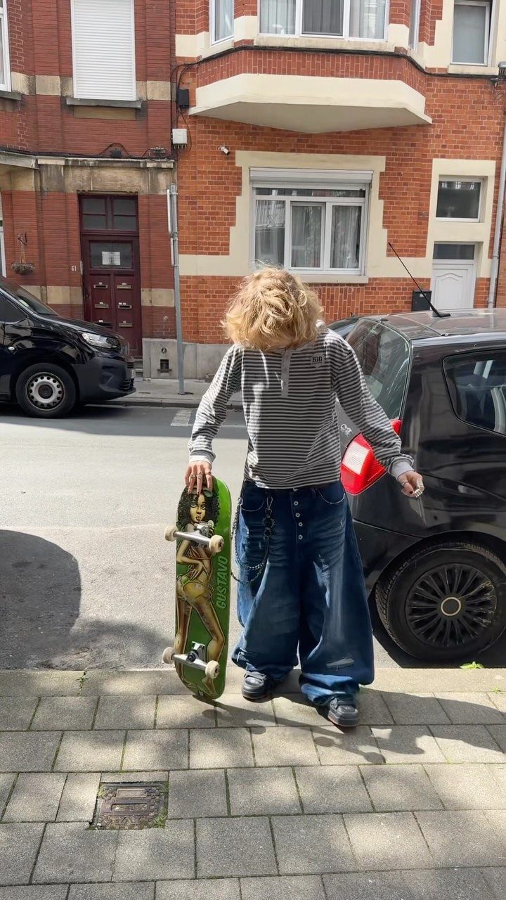 Person in baggy jeans and striped shirt carrying a green skateboard on a city sidewalk.