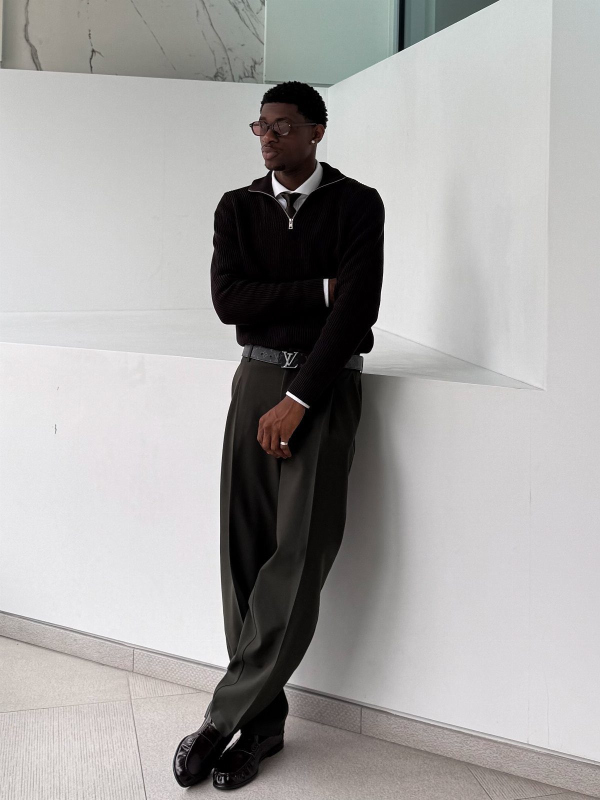 A man in smart-casual attire leaning against a white wall in a minimalist environment.
