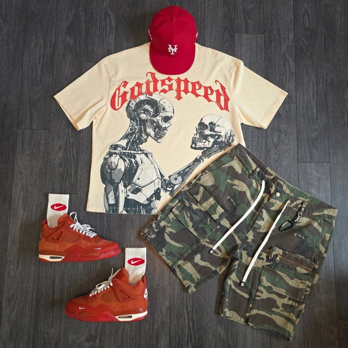 Streetwear outfit featuring a graphic t-shirt, camo shorts, red cap, and red sneakers laid on dark wood.