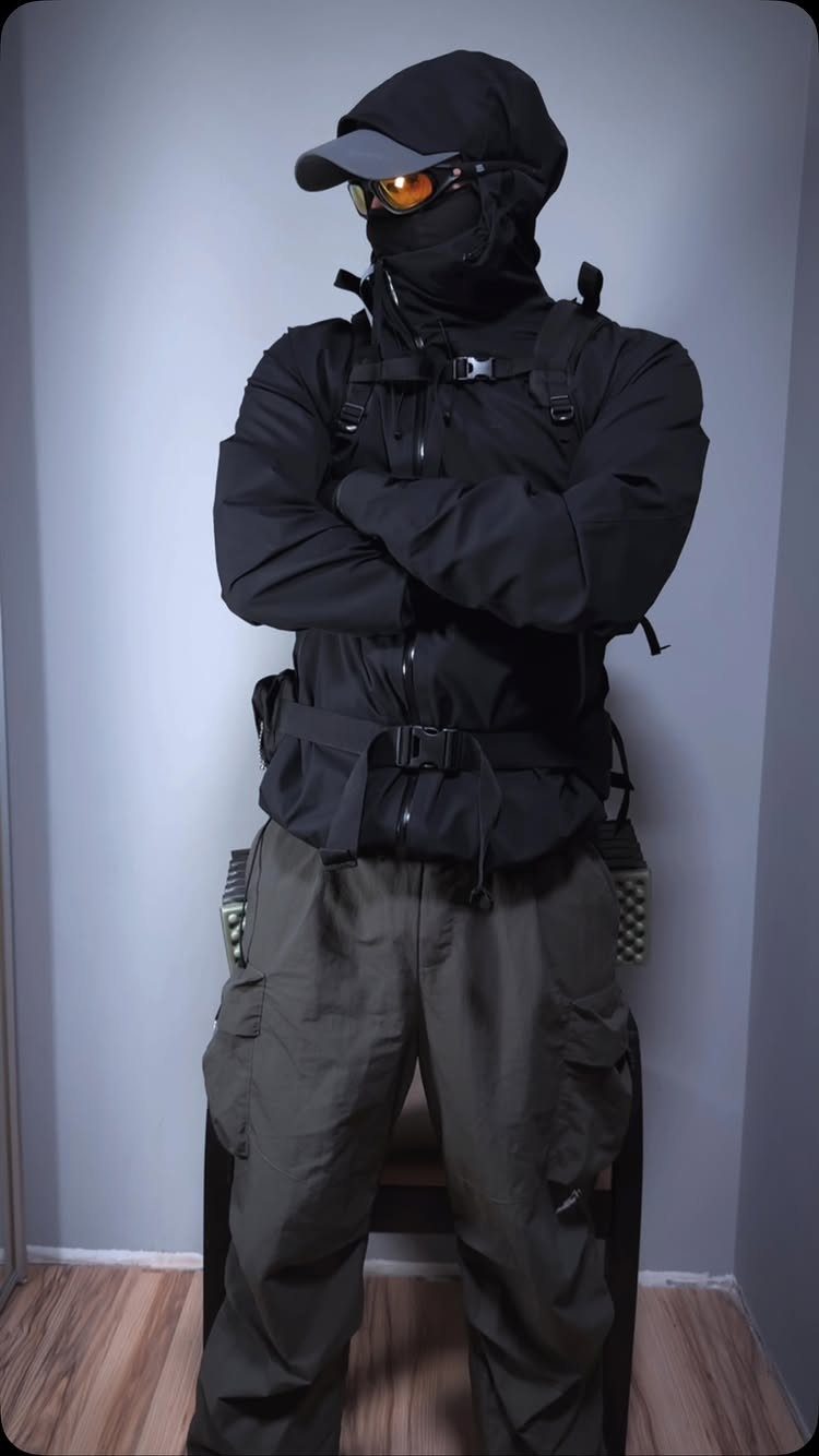 A person dressed in black tactical gear and olive cargo pants standing with arms crossed.