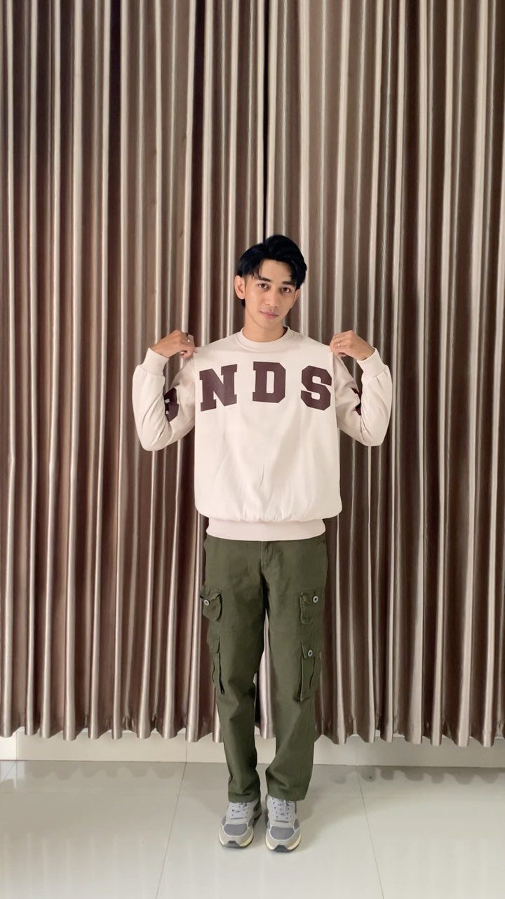 Young man wearing a cream sweatshirt and olive green cargo pants standing before tan curtains.