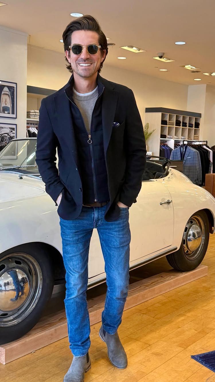 Smiling man in a navy blazer, blue jeans, and grey boots standing by a white car.
