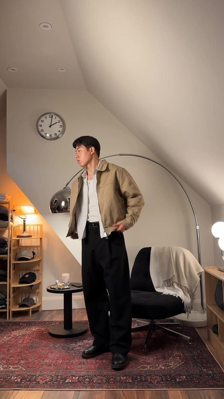 A man in a tan jacket, white shirt, and black trousers standing in a room.