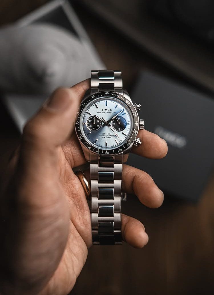 A hand holding a silver Timex chronograph watch with a light blue dial.