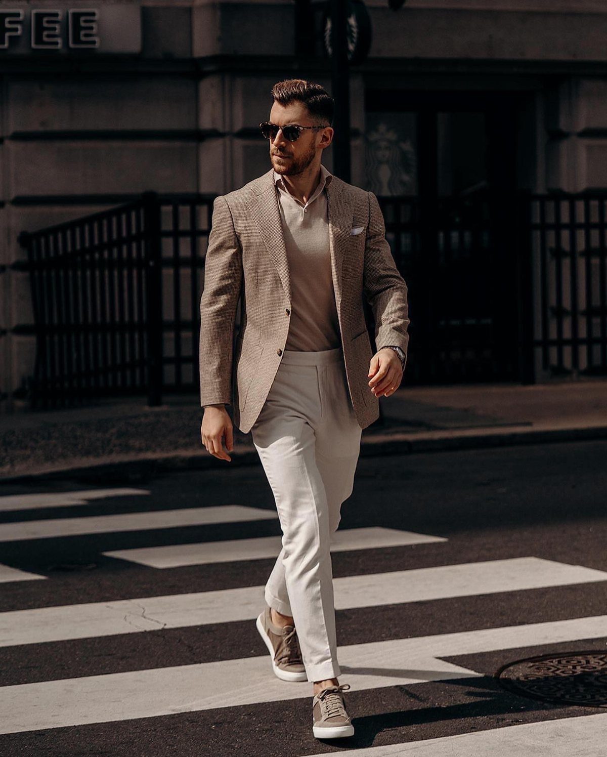 Man in a tan sport coat and white trousers walking across a city street crosswalk.