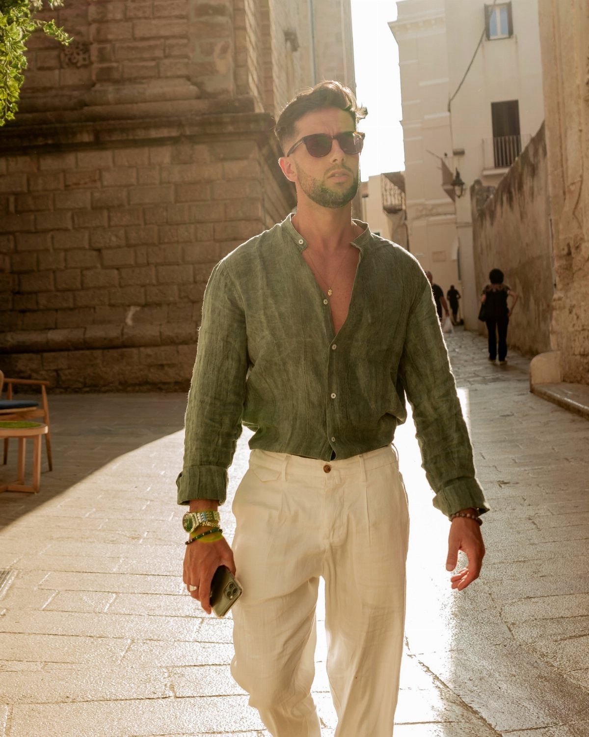 A man in a green linen shirt and white trousers walking through a sunny European street.