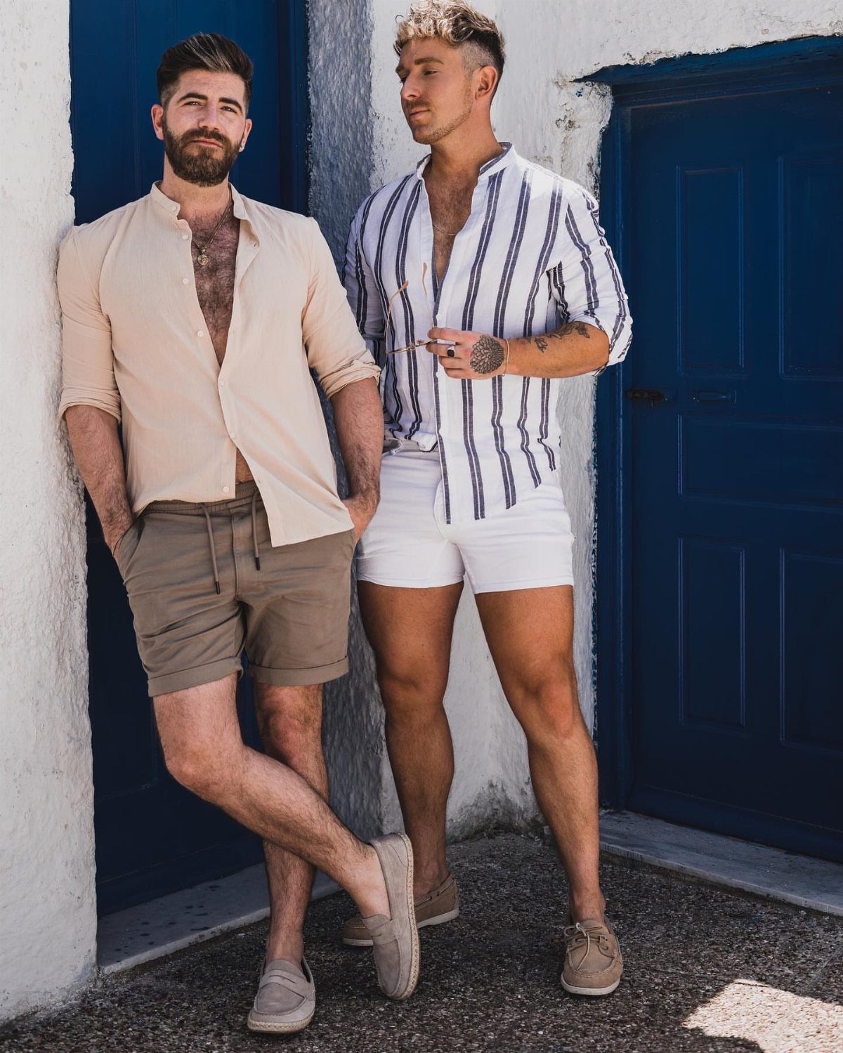 Two men posing in casual summer outfits against a white wall and blue doors.