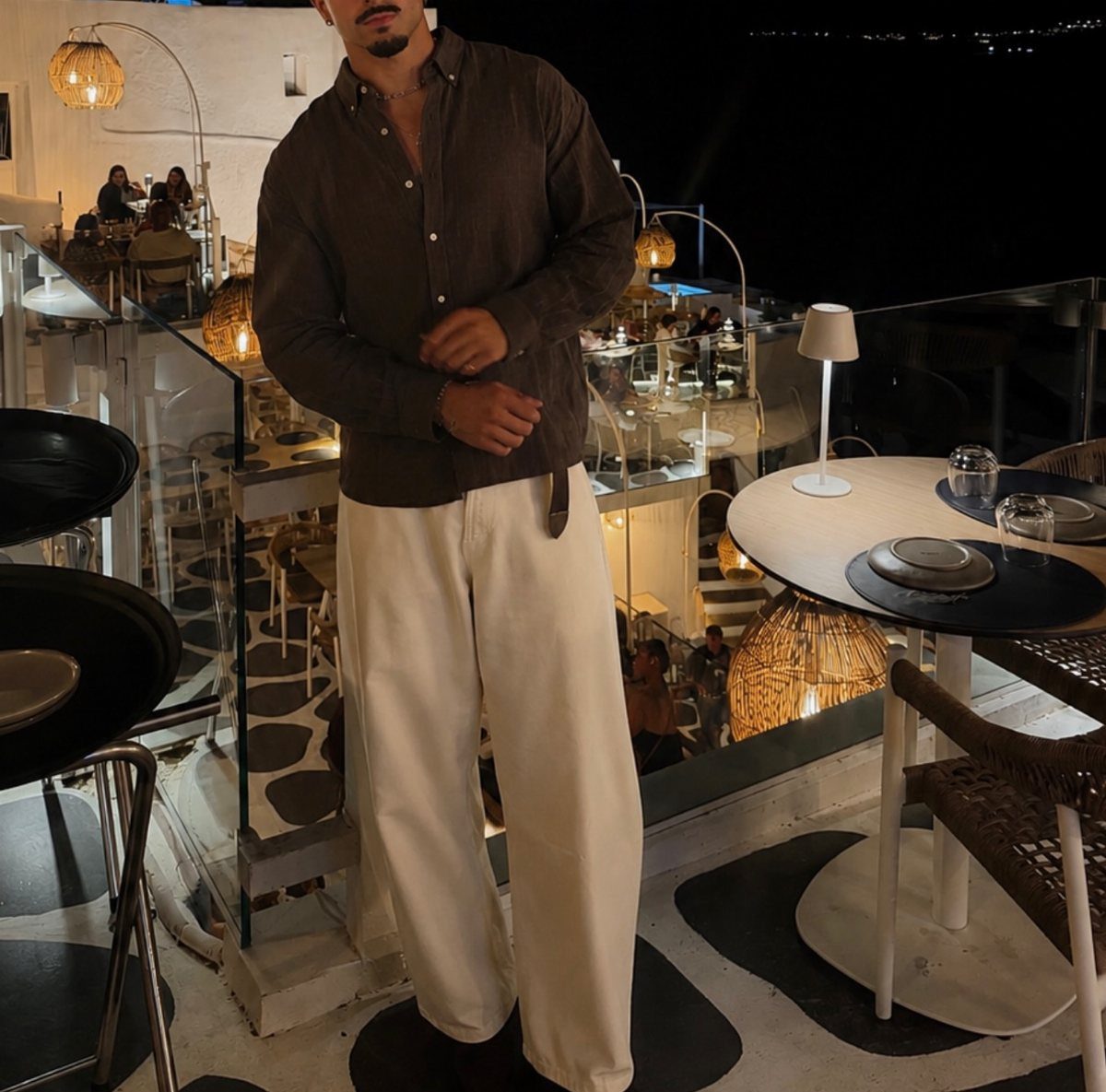 Man wearing a brown button-up shirt and cream trousers standing on a balcony at night.