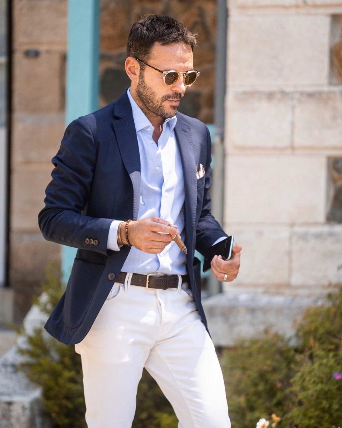 A man wearing a navy blue blazer, light blue shirt, and white trousers outdoors.