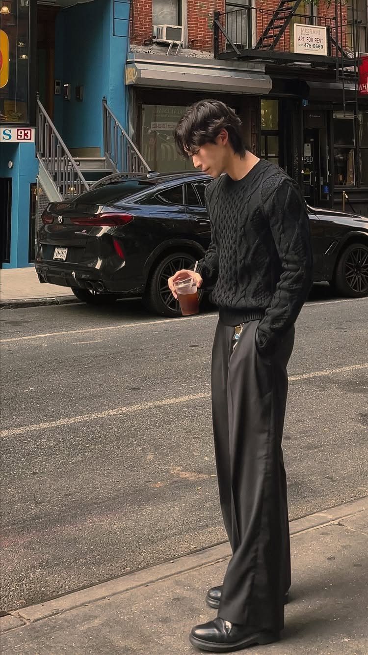A young man in an all-black textured sweater and wide-leg trousers standing on a city street.