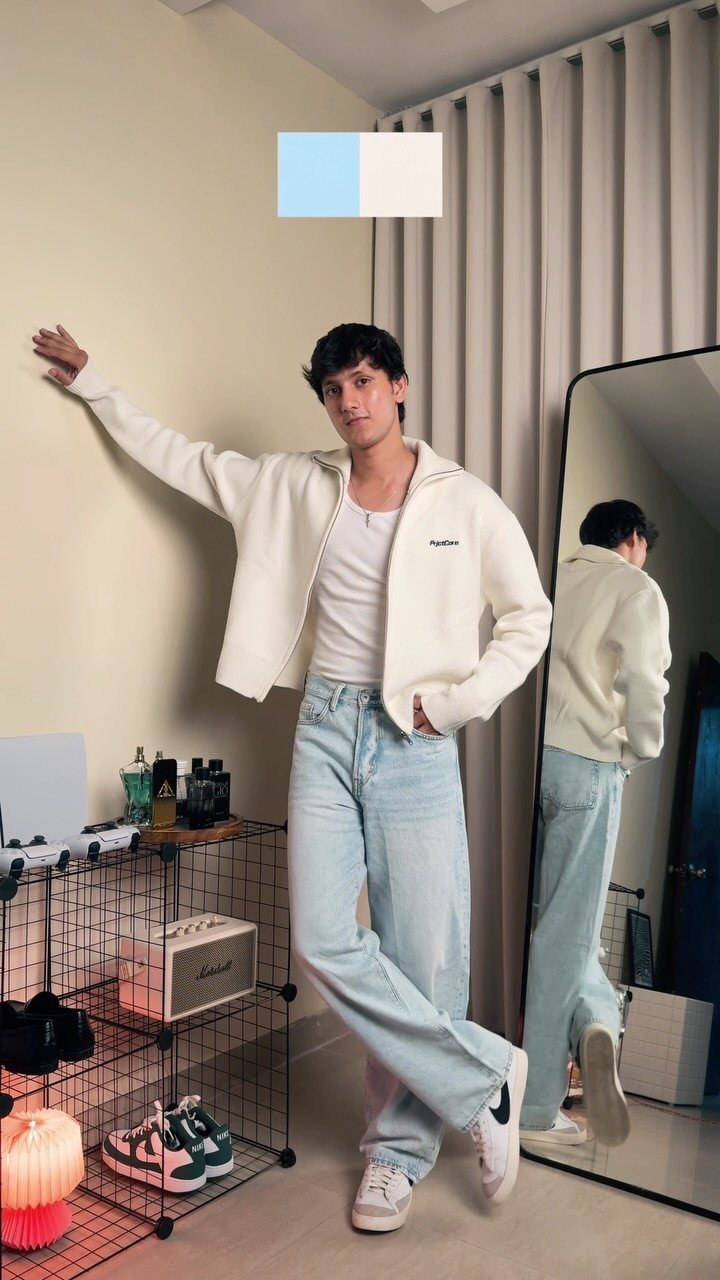 A young man posing in light blue jeans, a white jacket, and white sneakers.