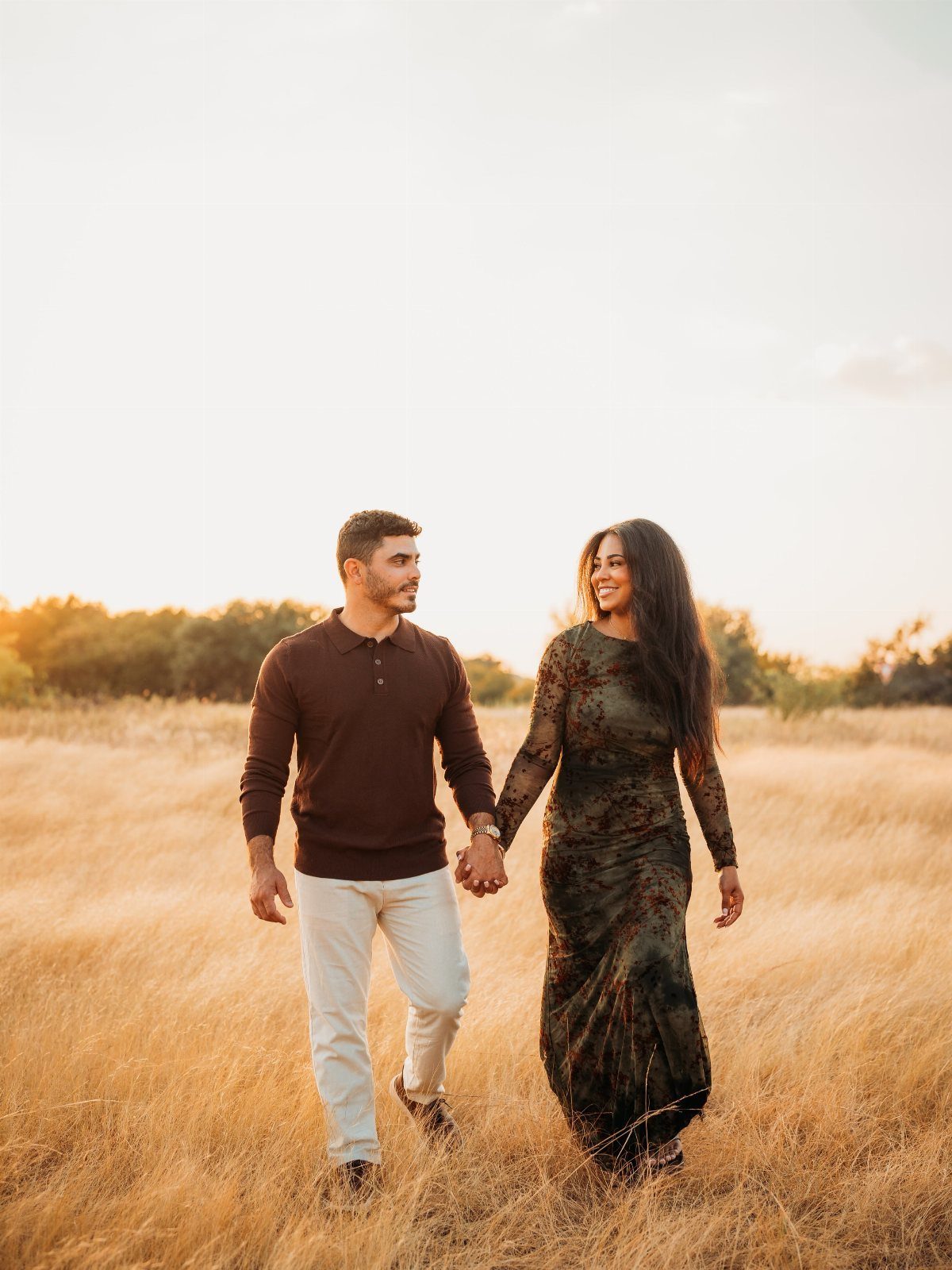 A man and woman walking through a golden field at sunset holding hands.