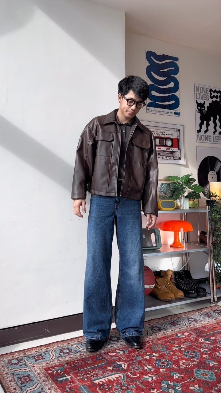 A man wearing a brown leather jacket, wide-leg jeans, and black boots standing on a rug.