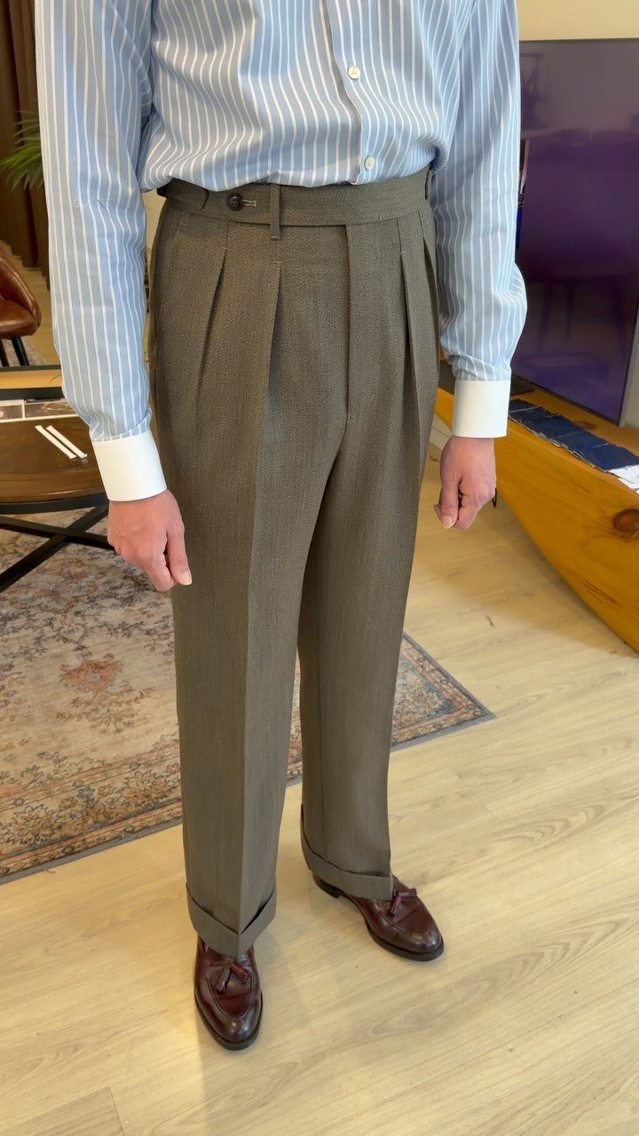 A person wearing grey pleated tailored trousers with cuffs and a blue striped shirt.