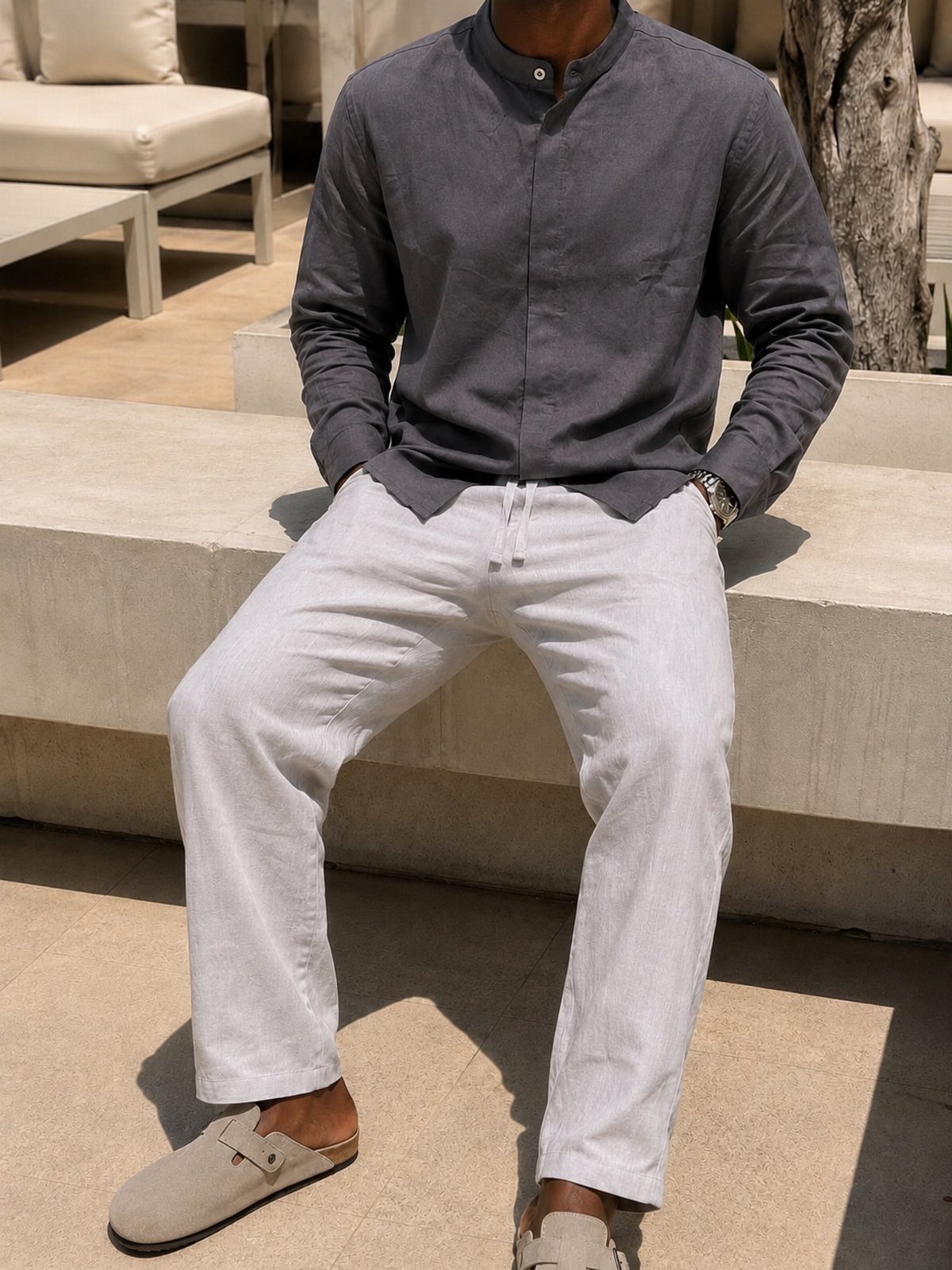 Man sitting outdoors wearing a dark grey linen shirt and light grey linen trousers with beige loafers.