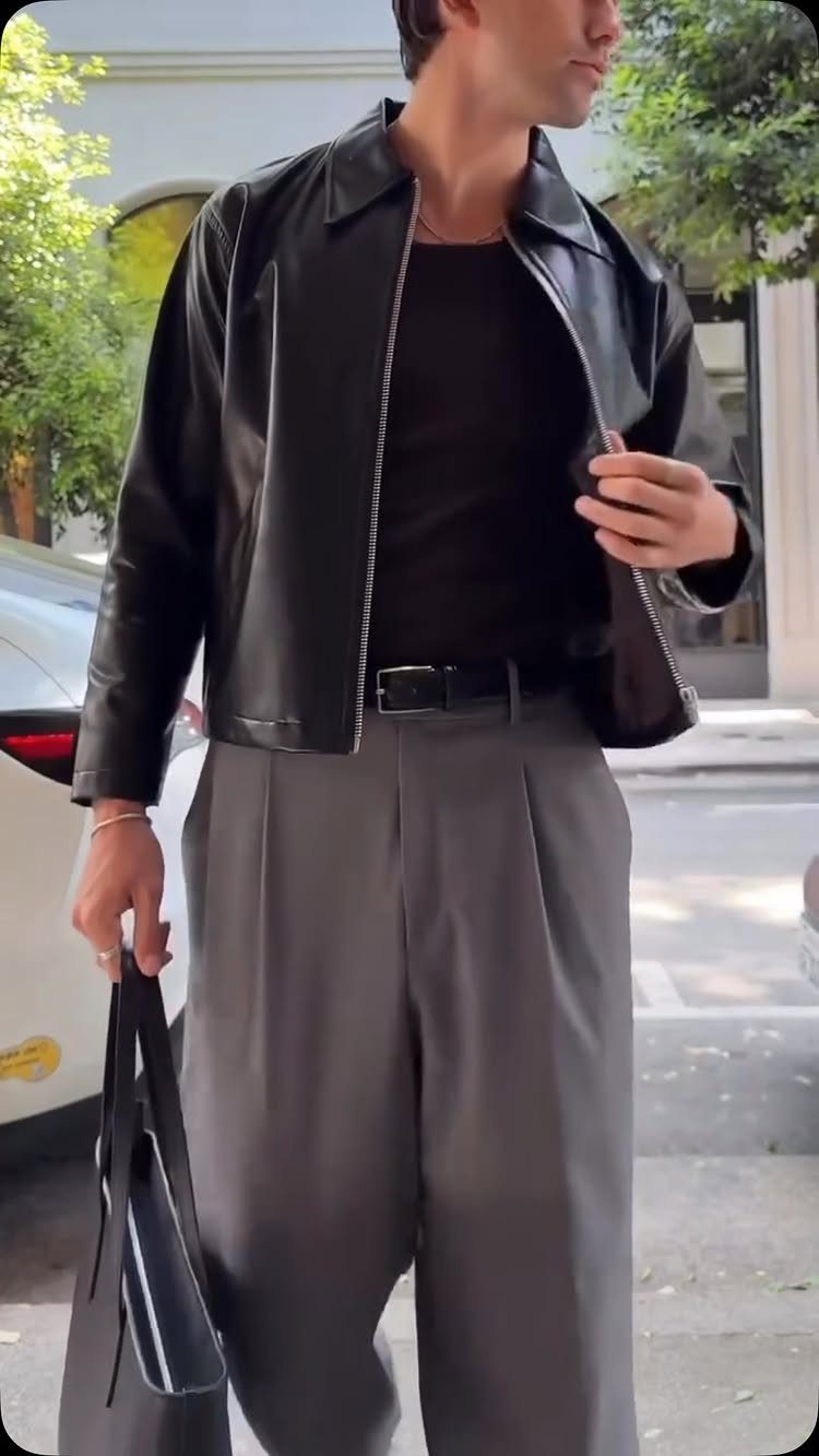 A person wearing a black leather jacket, black t-shirt, and grey trousers holding a black bag.