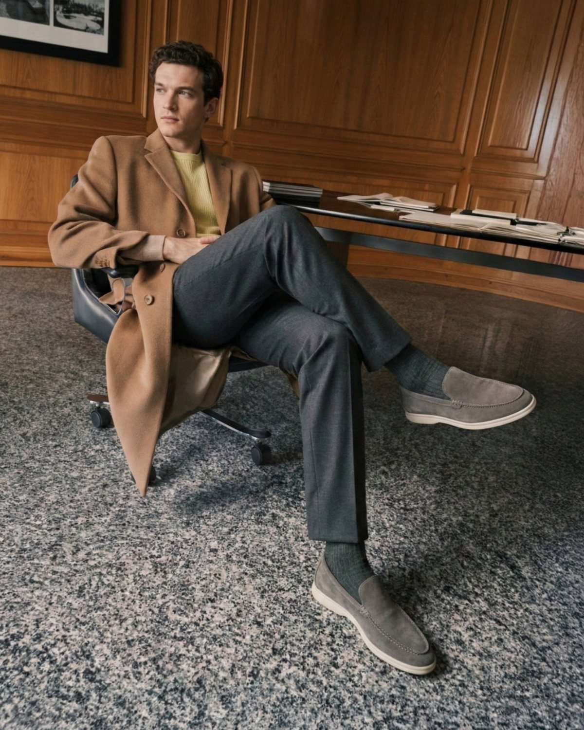 A man sitting in an office chair wearing a tan coat and grey trousers.