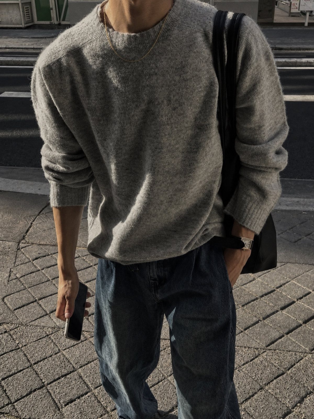Man wearing a grey crewneck sweater and blue jeans standing on a paved street.