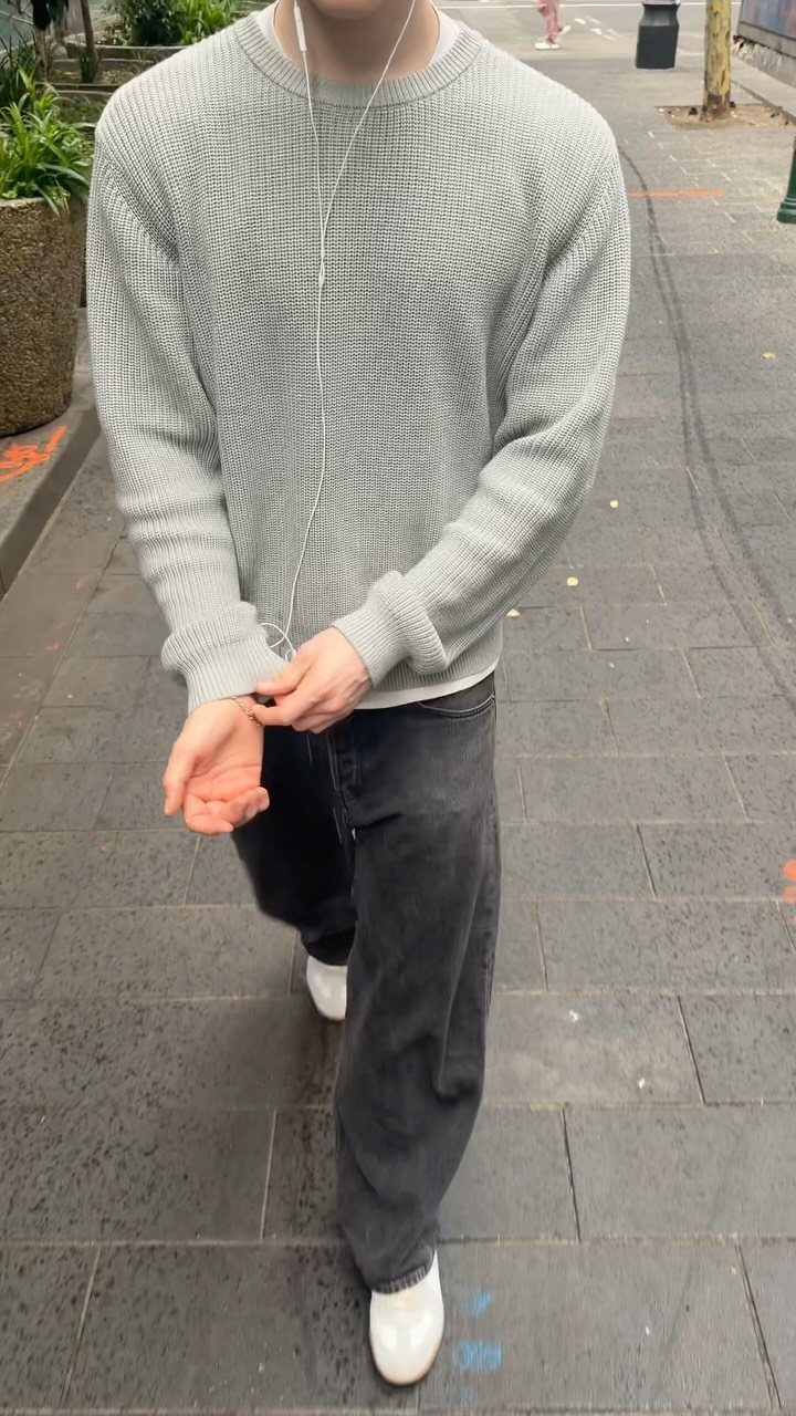 Man wearing a light grey textured sweater, black jeans, and white sneakers on a sidewalk.