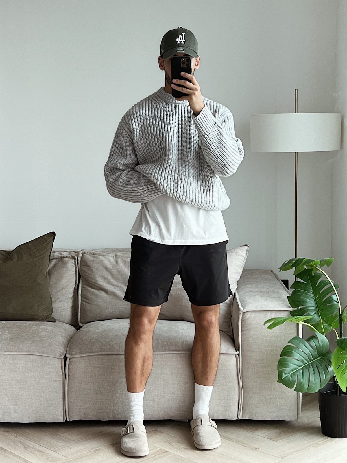 Man in a grey ribbed sweater, white t-shirt, black shorts, and beige clogs standing in a living room.