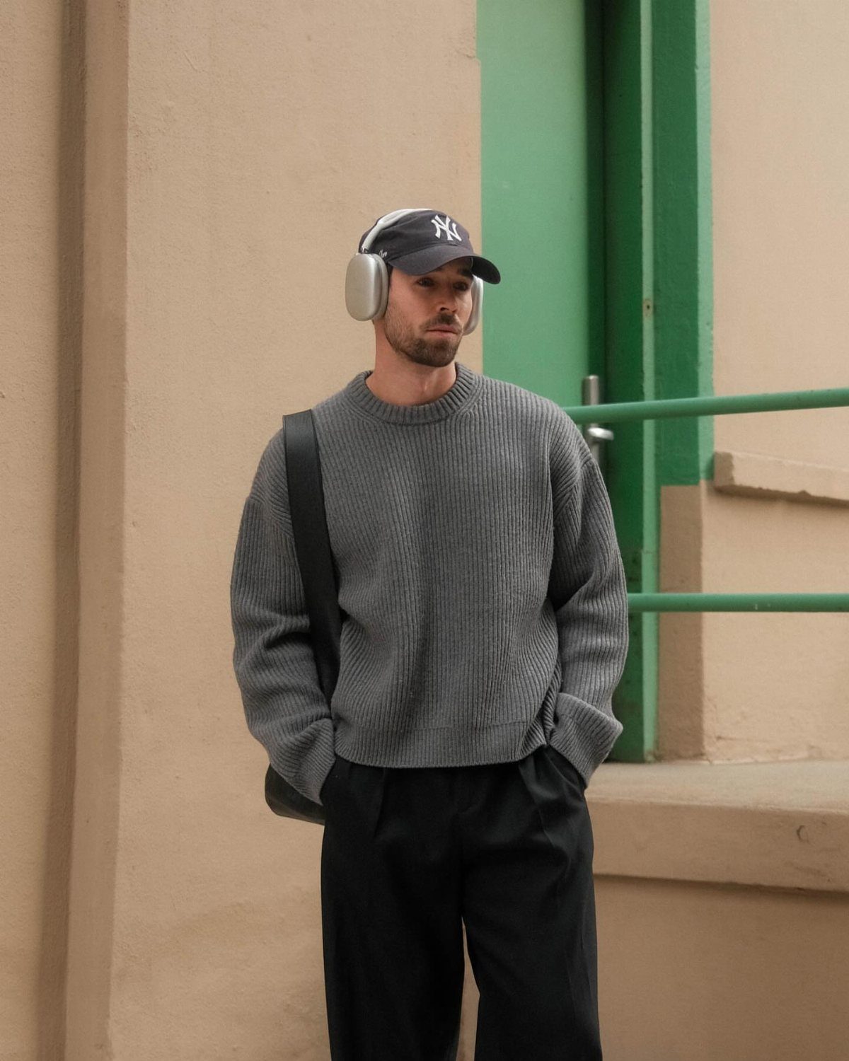 Man in a grey ribbed sweater, black trousers, and earmuffs standing by a building.