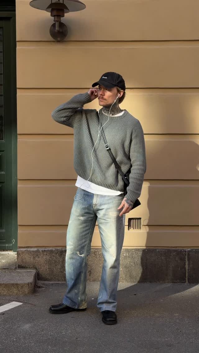 A man wearing a grey ribbed sweater, light blue jeans, and a black cap standing outdoors.